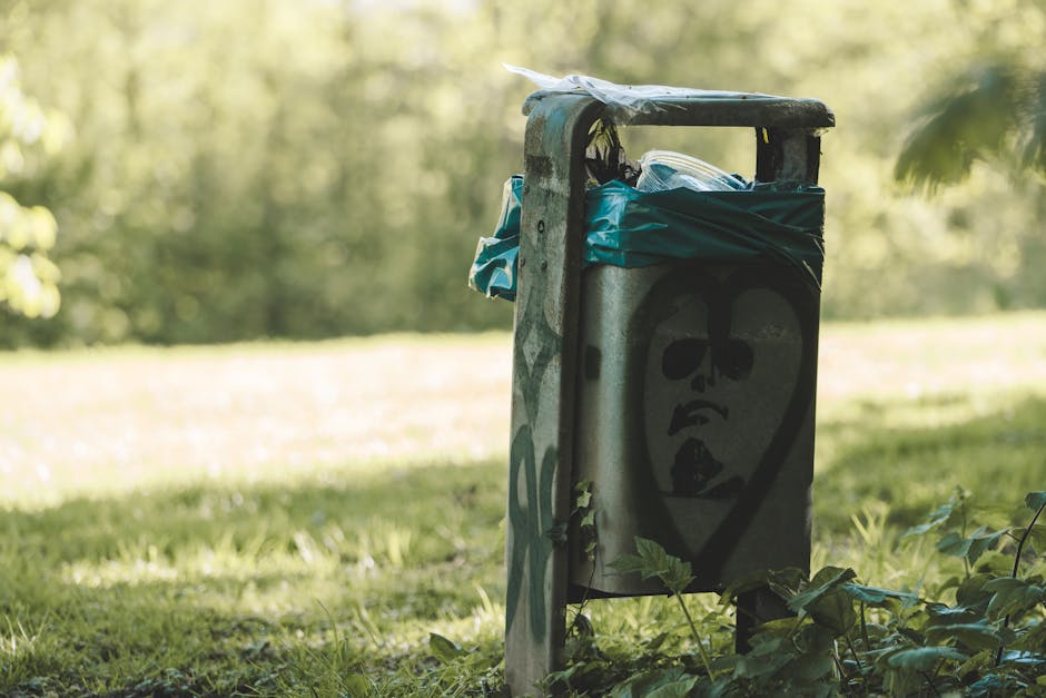 A weathered metal waste bin located in a garden area, partially covered with a blue plastic bag and some clear plastic wrapping on top. The bin features a black graffiti-style face with large eyes, a
