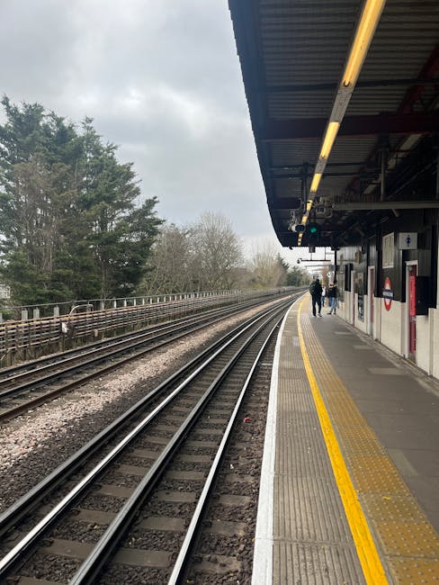 The image depicts a train platform at Mill Hill Broadway station, with railway tracks running parallel along the left side. The platform surface is made of concrete, featuring a tactile yellow warning