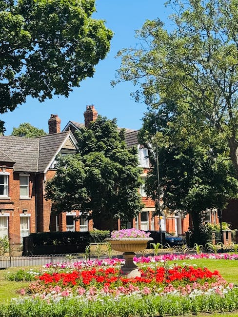 A picturesque urban garden scene featuring a neatly maintained lawn bordered by vibrant flower beds with red, pink, and white blooms. In the center, a classical stone planter filled with pink flowerin