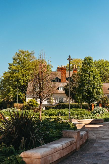 A picturesque urban garden scene featuring a neatly maintained lawn bordered by vibrant flower beds with red, pink, and white blooms. In the center, a classical stone planter filled with pink flowerin